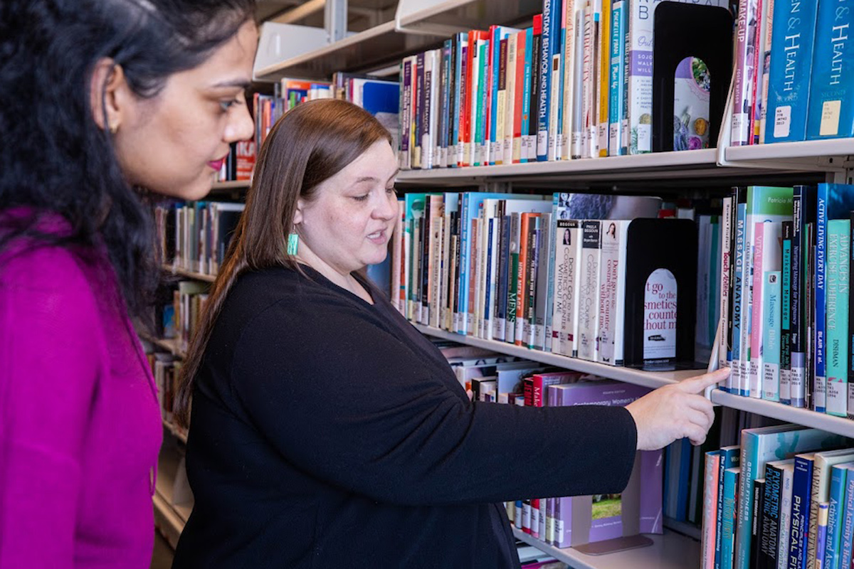 A Library team member pointing to a book in the Library stacks for a student standing behind them.