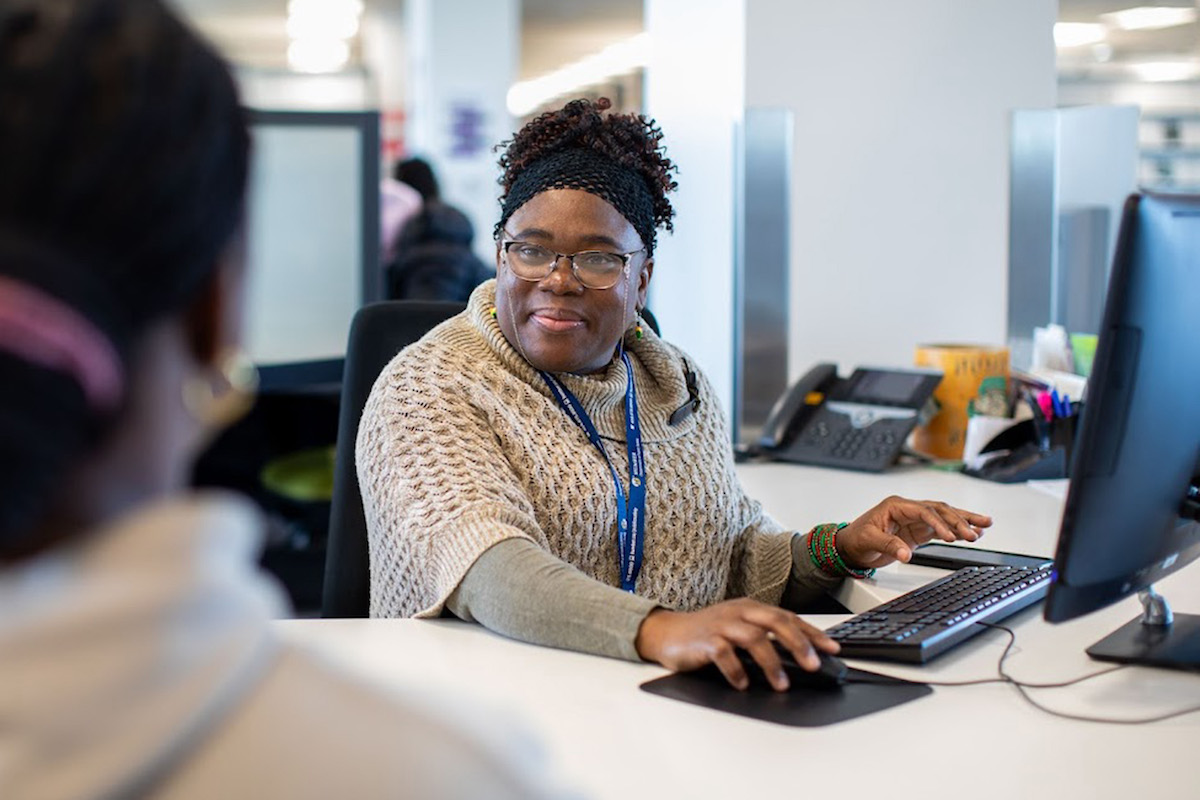 A smiling Library team member seated behind a Library desk typing on a computer while facing a student.