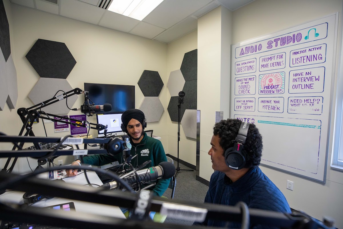 Two students sitting in an audio recording studio, wearing headphones and speaking into microphones.