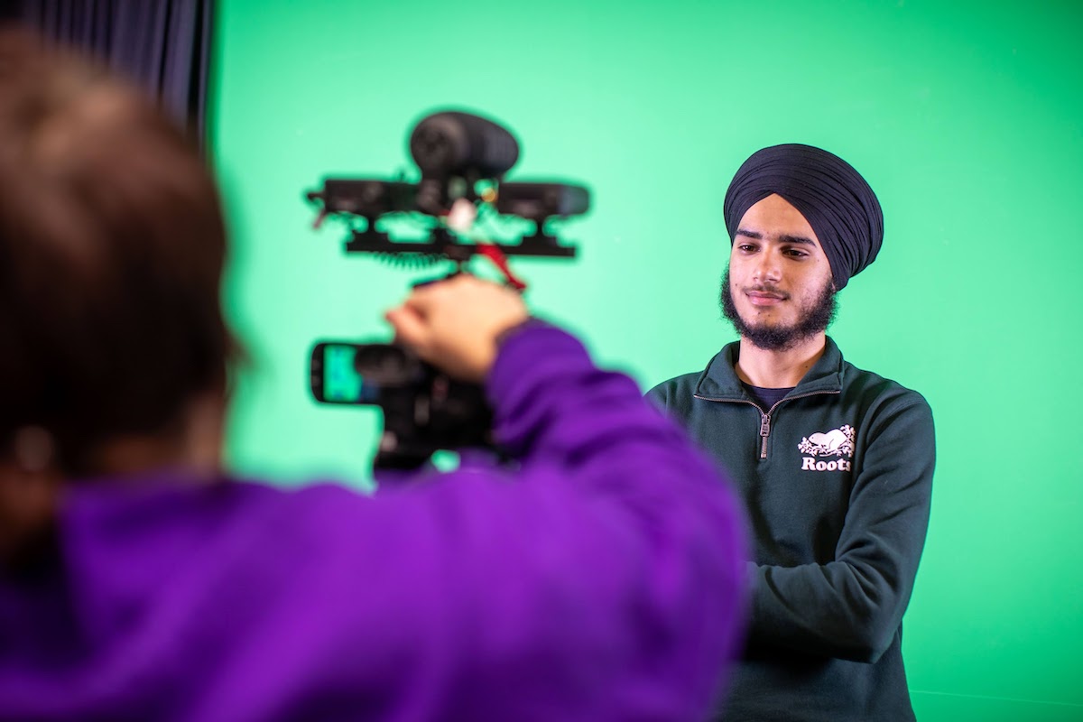 A student adjusting a camera on a tripod, while another student stands in front of a green screen, smiling, with crossed arms.