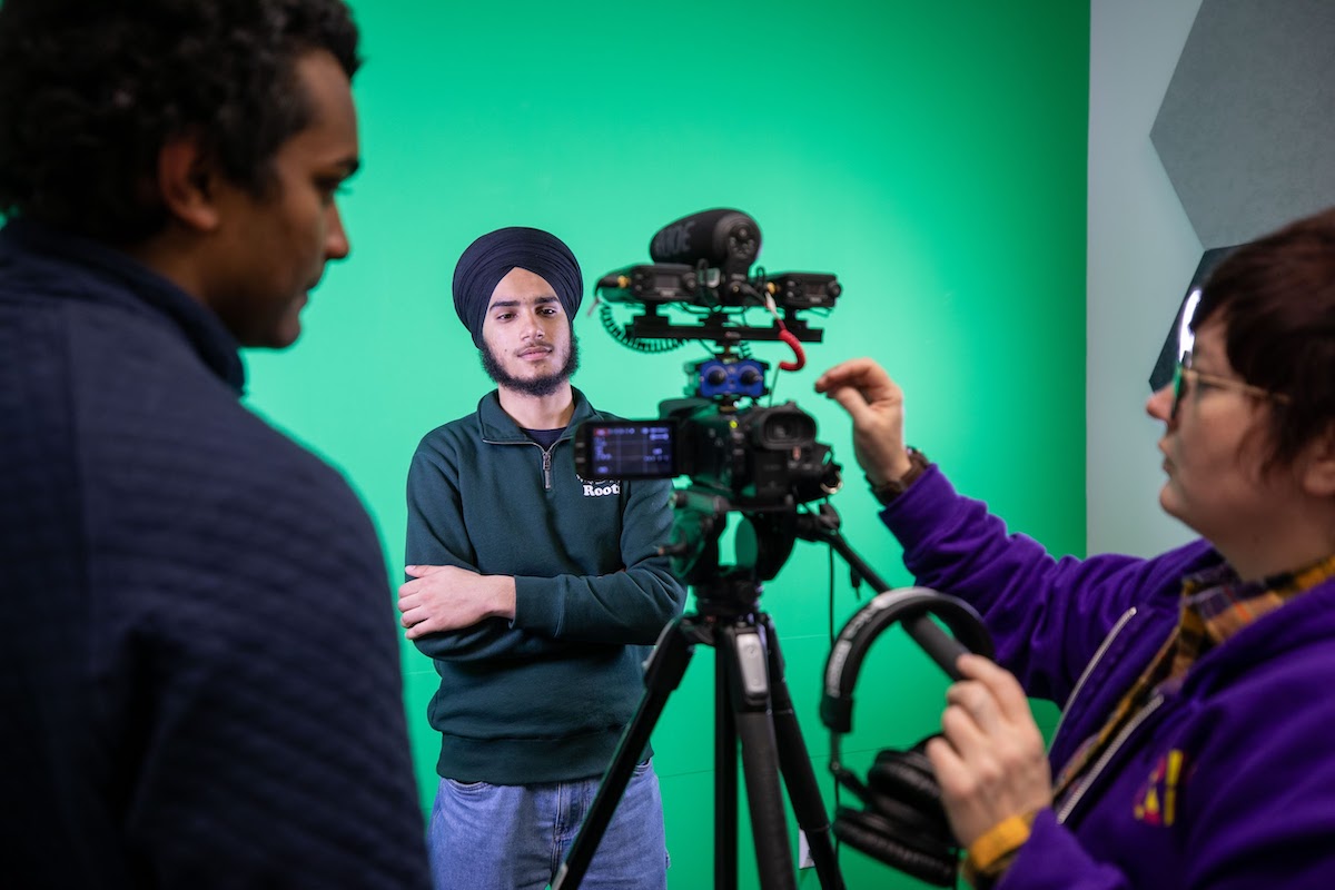 Two students adjusting a camera on a tripod, while a third student stands in front of a green screen, smiling, with crossed arms.