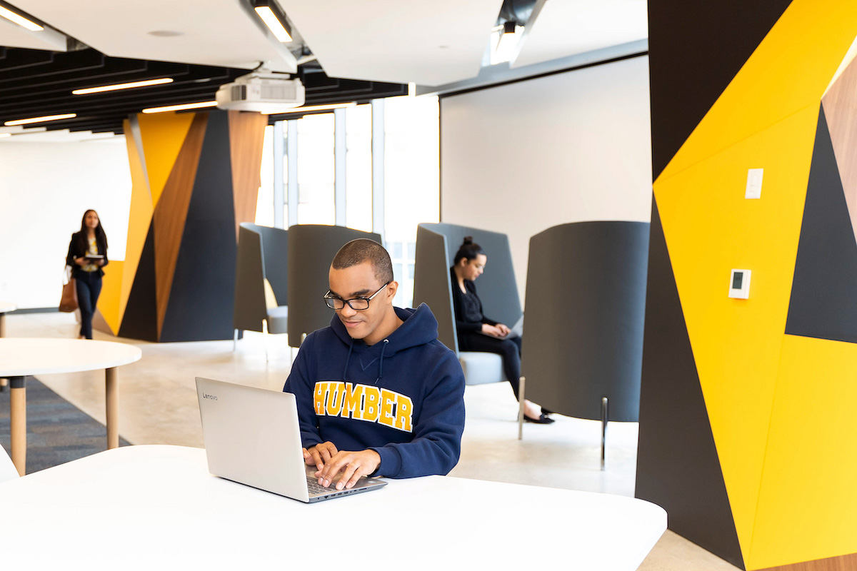 A student seated at a table typing on a laptop. In the background, a student is sitting on the grey soft seats and another is walking by.