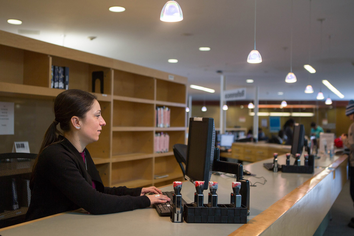 A Library team member staffing a Library desk. They are seated at a computer workstation and typing.