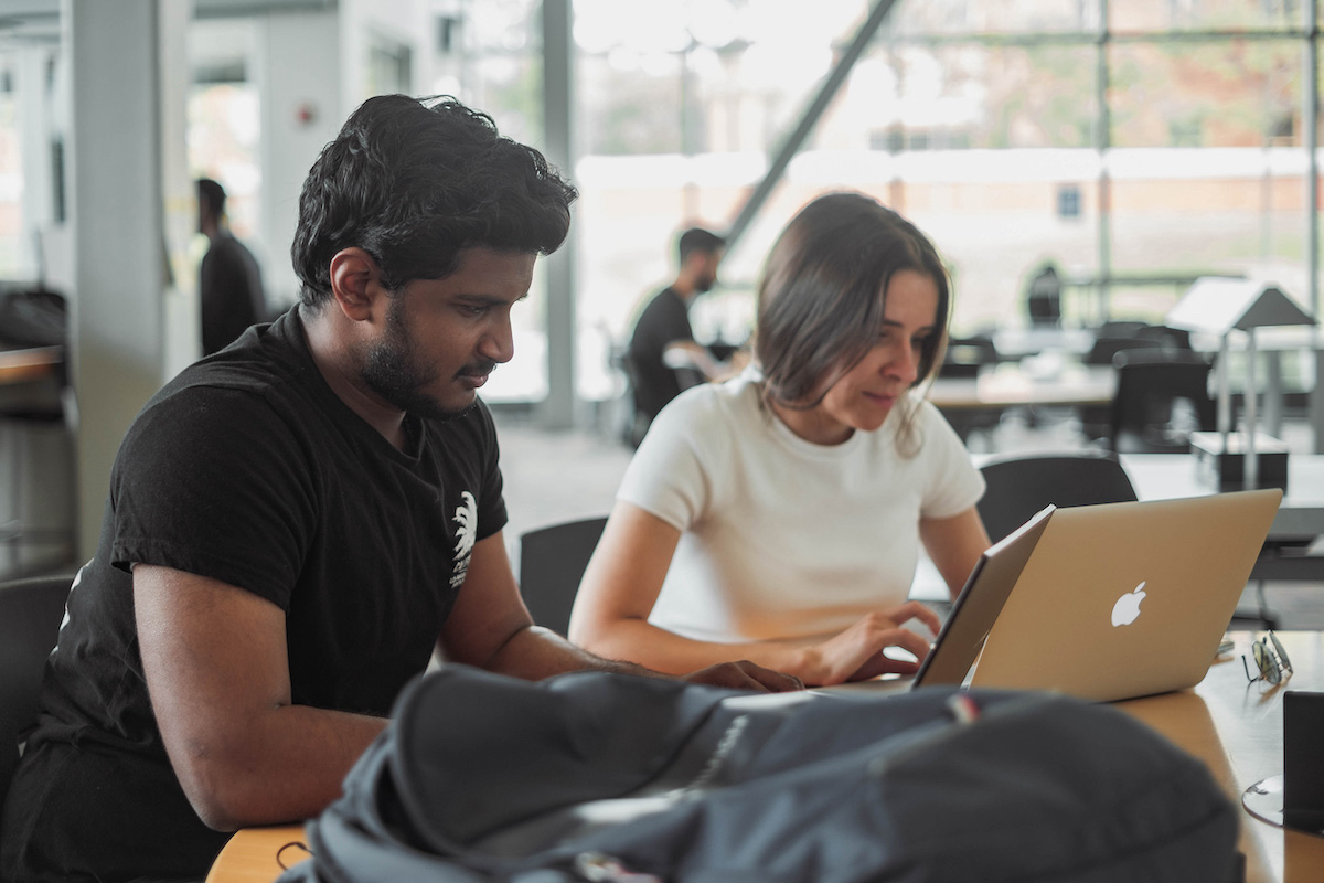 Two students sitting side by side in a sunny, open-concept space, typing on their laptops.