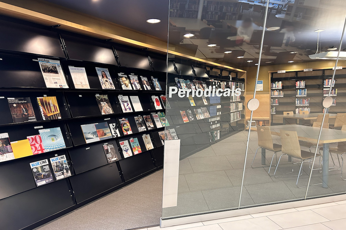 Enclosed Periodicals area that has wall shelves with magazines and a table and chairs.