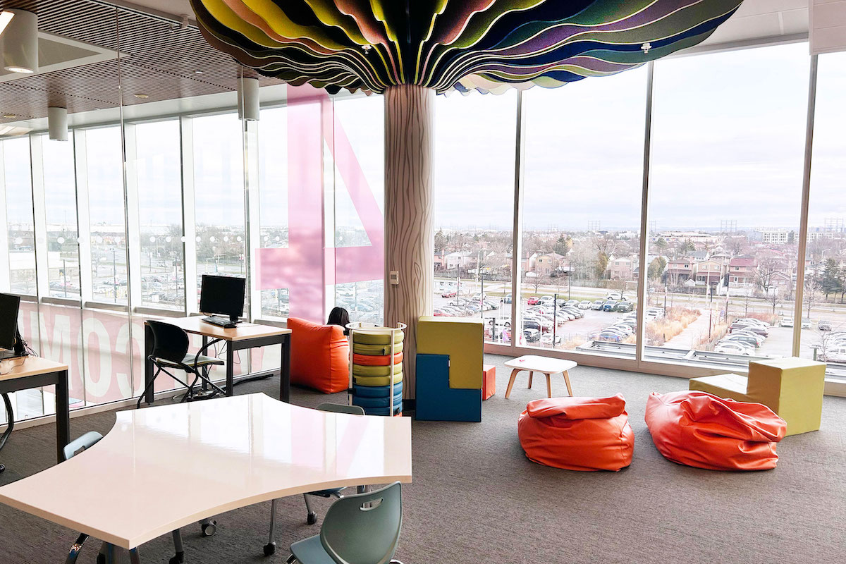 A cheerful Library seating area for children and adults, with a multi-coloured modular sofa, tables and orange bean bag chairs. A structural column is decorated to look like a tree.