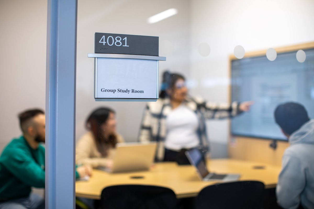 Study room with three students seated at table looking at a standing student pointing to a whiteboard.