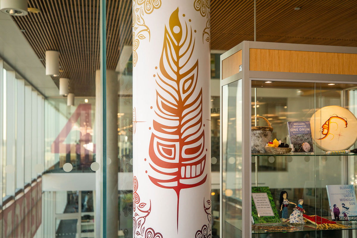 A pillar in Humber’s North Library, painted with a red feather design by Ojibwe artist Patrick Hunter. Beside the pillar, a glass cabinet displays small cultural objects such as art, dolls and carvings.