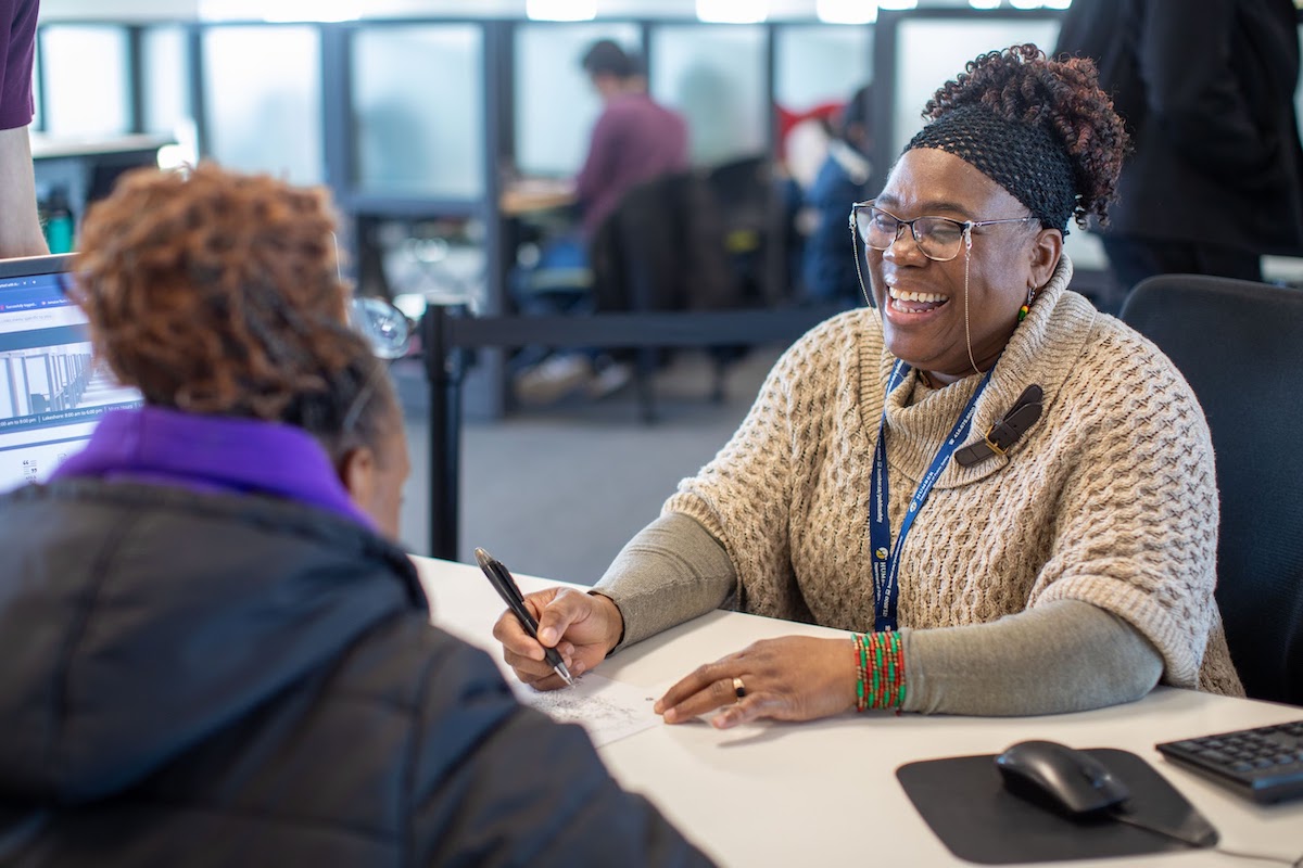 A laughing Library team member seated behind a Library desk, writing something down on a piece of paper for a Library visitor.