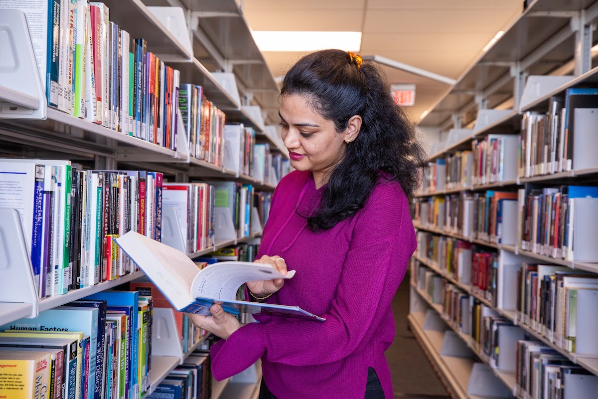 A smiling student standing in the Library stacks and flipping through a hardcover book.